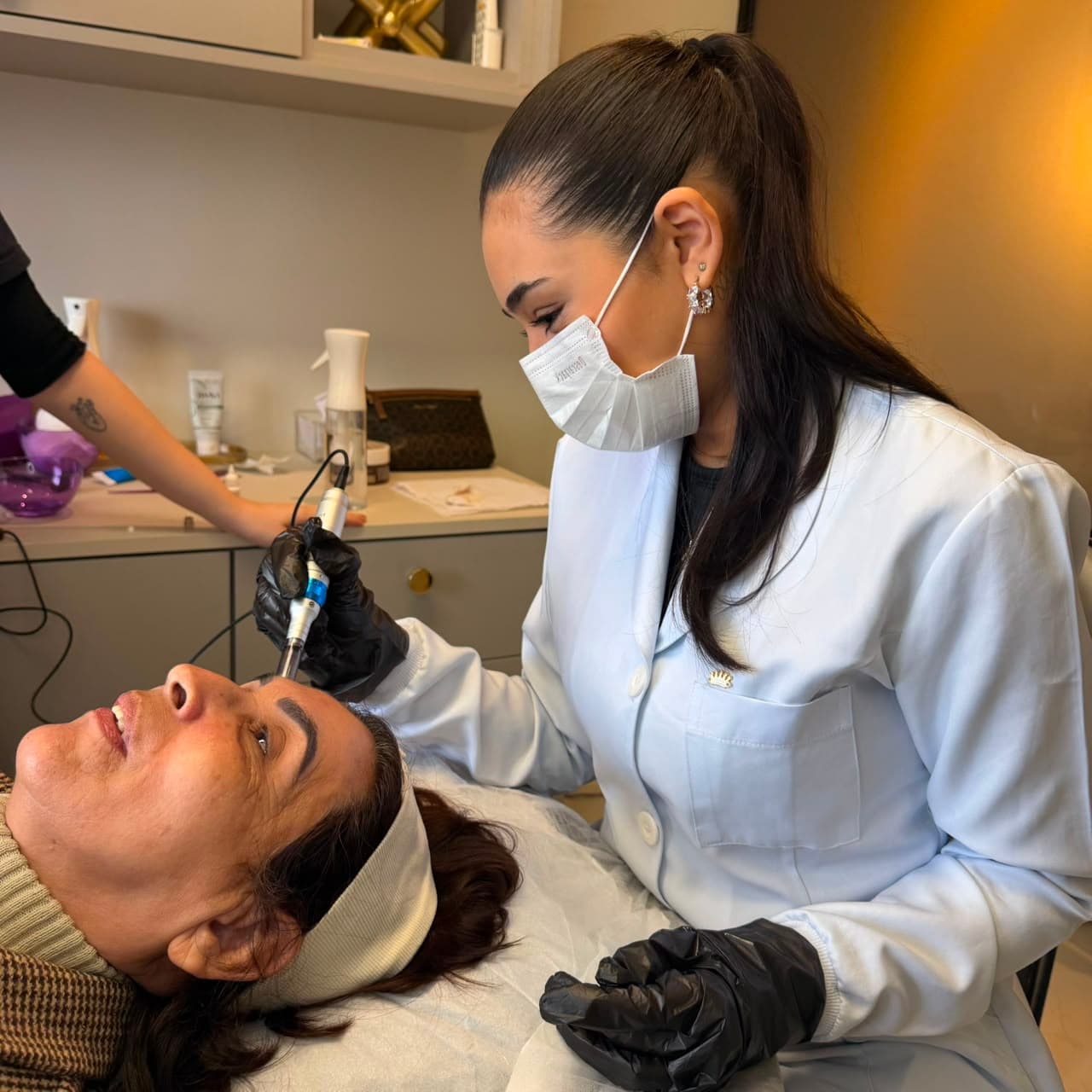 Aluna do SkinLab aplicando microagulhamento na testa de uma paciente, de jaleco, máscara e luvas.