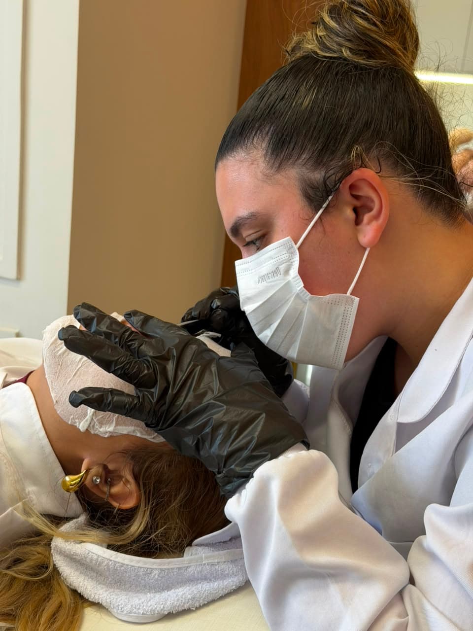 Aluna do SkinLab fazendo a extração na testa de uma paciente, de luvas pretas e máscara.